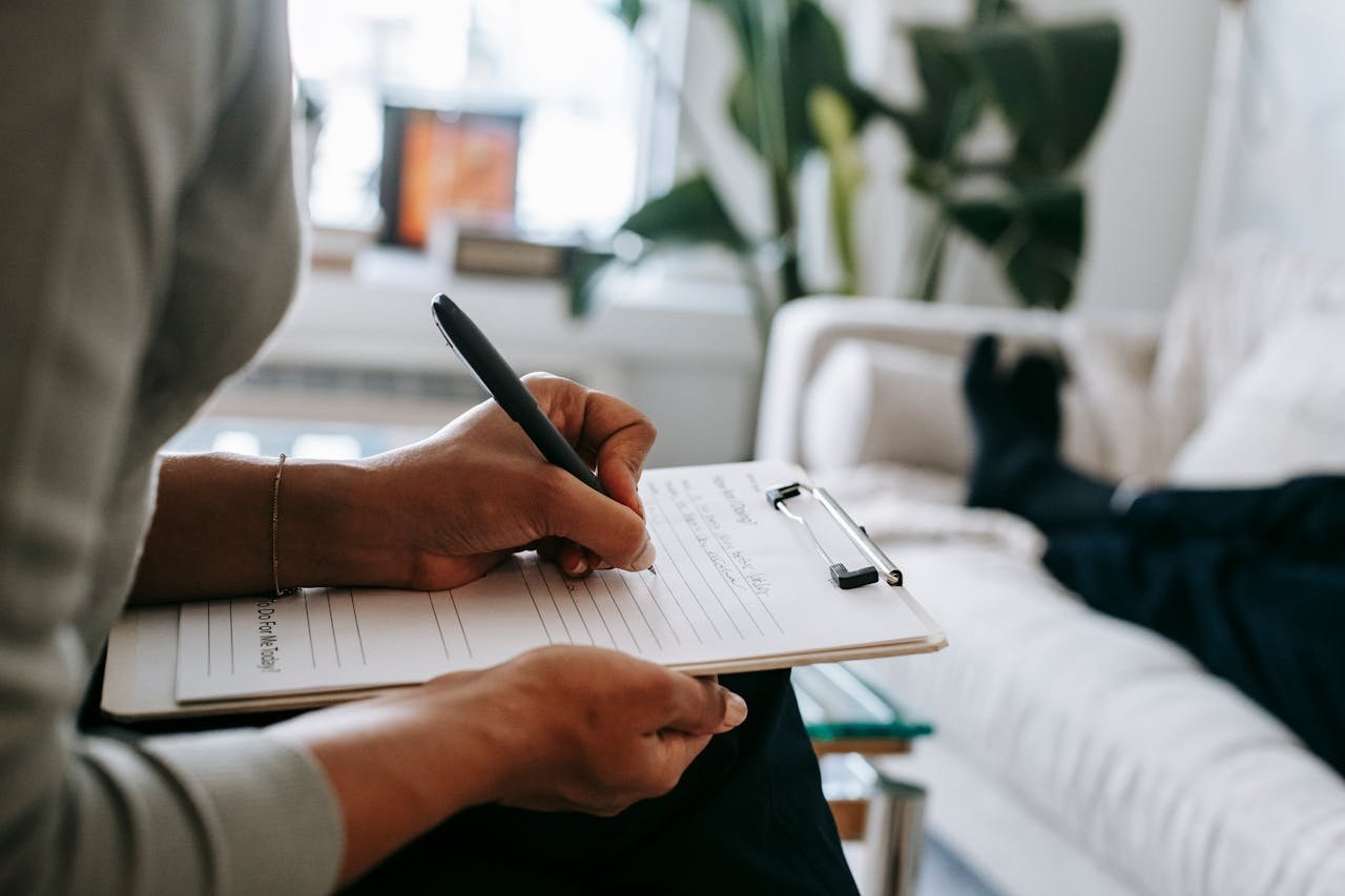 services-01 Unrecognizable ethnic female therapist taking notes on clipboard while filling out form during psychological appointment with anonymous client lying on blurred background